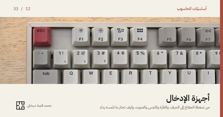 غلاف مقالة أجهزة الإدخال: صورةٌ مقرّبة للزاوية العليا من لوحة مفاتيح ميكانيكيّة بأزرارٍ رماديّة فاتحة، وزرّ Esc أحمر في طرفها، على مكتبٍ خشبيّ. وفوق الصورة اسم السلسلة «أساسيّات الحاسوب» والرقم 12 من 33، وتحتها العنوان «أجهزة الإدخال» وسطرٌ يقول: من ضغطة المفتاح إلى الحرف، والفأرة واللمس والصوت، وكيف تختار ما تلمسه يدك.