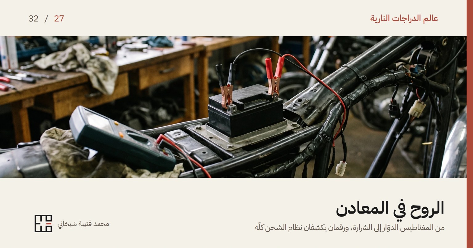 بطارية دراجة نارية في مهدها والمقعد مرفوع، وعلى قطبيها ملقطا مقياس متعدّد، أحدهما بمقبض أسود والآخر أحمر، ويمتدّ سلكاهما إلى مقياس راقد على خرقة إلى اليسار وشاشته مائلة لا يُقرأ عليها شيء. وحول البطارية أنابيب الهيكل وضفيرة أسلاك ملفوفة ووصلات بيضاء، وخلفها منضدة ورشة ومفاتيح معلّقة على الجدار.
