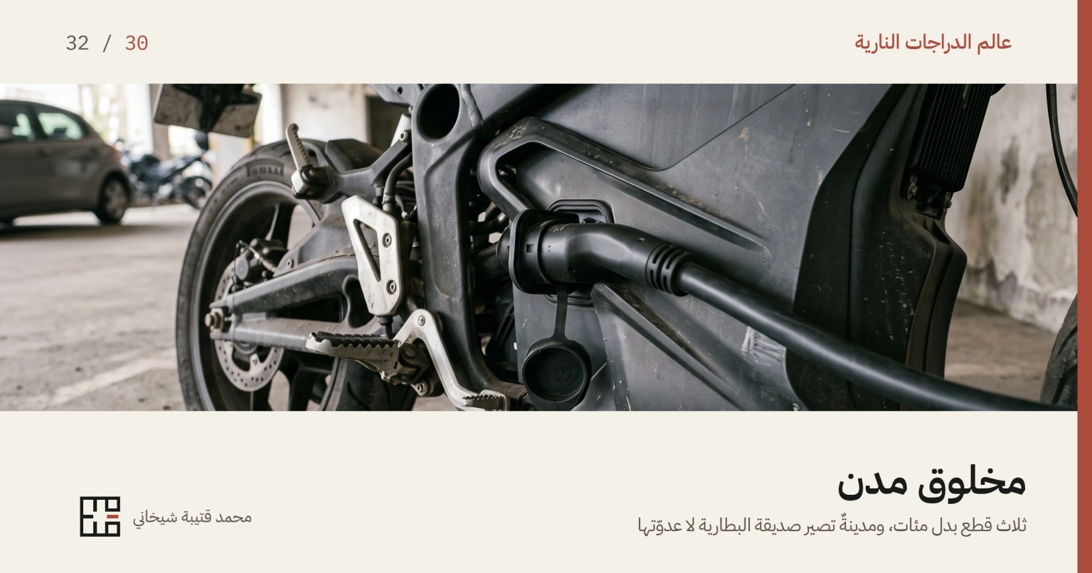 مقبس شحن مفتوح في جانب دراجة نارية كهربائية وقابس غليظ مغروز فيه، والكابل يمتدّ خارج الكادر، وحول المقبس هيكل الدراجة ومسند القدم والعجلة الخلفية وقرص فرامل، في مرأب خرسانيّ.