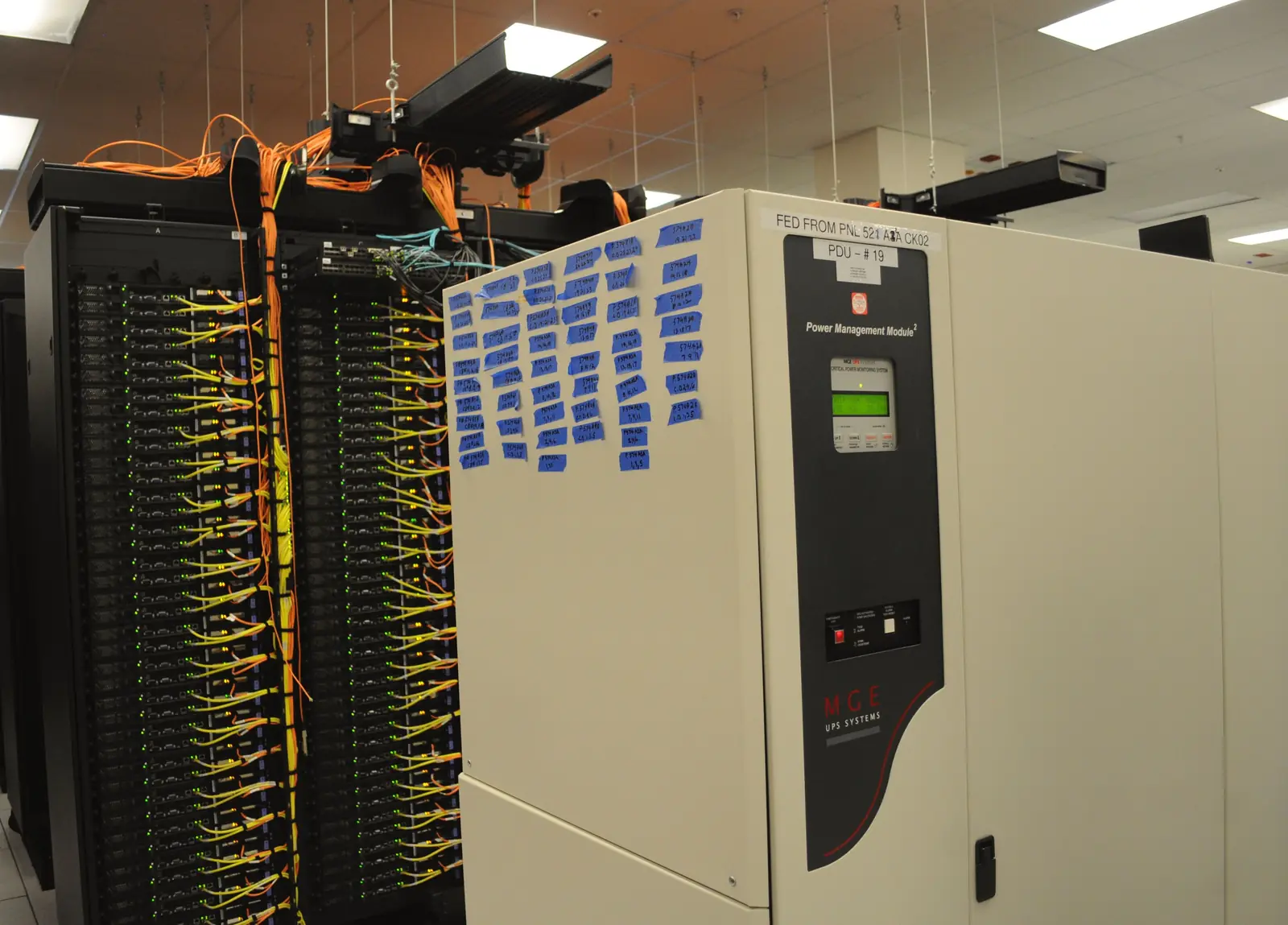 خزانةٌ عاجيّة اللون كبيرة عليها شاشةٌ خضراء صغيرة ولافتة «Power Management Module» وشعار MGE UPS Systems، وعلى جانبها عشرات البطاقات الزرقاء المكتوبة بخطّ اليد، وإلى يسارها خزانتا خوادم سوداوان تتدلّى منهما كابلاتٌ صفراء وبرتقاليّة، وفوقها مساراتٌ معلّقة للكابلات.